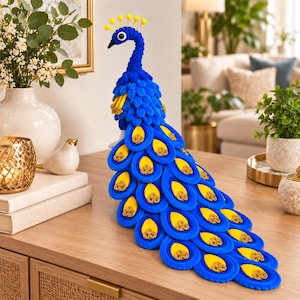 May include: A decorative peacock figurine crafted from blue felt with a long, cascading tail. The tail features layered, oval-shaped feathers in blue and yellow, each accented with a gold spiral detail. The head has yellow pom-pom crests.