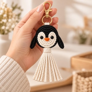 May include: A macrame keychain featuring a penguin design with a black and cream cord construction. The penguin has black side wings, a cream face, two black bead eyes, and an orange beak. A gold-toned metal lobster clasp is attached to the top.