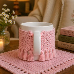 May include: A white ceramic mug featuring a light pink crocheted cozy with a textured star-like pattern and a row of small hanging tassels along the bottom edge. The cozy wraps around the mug, leaving the white handle exposed for easy gripping.