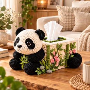 Könnte beinhalten: Ein rechteckiger Taschentuchbox-Überzug, gehäkelt in Form eines Pandas. Der Panda hat ein weißes Gesicht mit schwarzen Augenpartien, Ohren und Gliedmaßen. Die Box ist mit gehäkelten grünen Bambusstangen, kleinen rosa Blüten und grünen Blättern verziert.