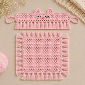 May include: A light pink crocheted cat-shaped headband with side loops and a matching square crocheted mat. Both items feature a textured stitch pattern and are finished with a decorative fringe of pink tassels along the bottom edges.