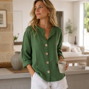 Puede incluir: Una camisa de manga larga en color verde oliva con cuello mao y cuatro botones circulares grandes de color claro en la parte delantera. La camisa se combina con pantalones cortos blancos y las mangas están remangadas hasta los codos.