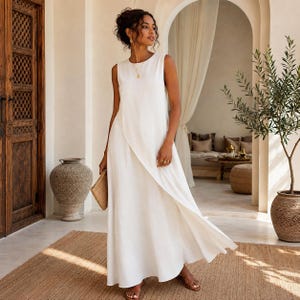 Könnte beinhalten: Ein ärmelloses, bodenlanges weißes Kleid mit Rundhalsausschnitt und einem überlappenden Vorderteil. Das Kleidungsstück besteht aus einem einfarbigen, blickdichten Stoff, der locker zum Saum hin fällt und eine schlichte Silhouette erzeugt.