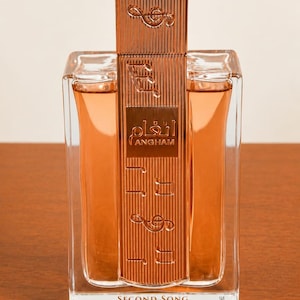 May include: A rectangular perfume bottle with a copper-colored cap and musical note accents. The bottle contains a light orange liquid and features the text "ANGHAM" in Arabic script. The base of the bottle reads "SECOND SONG".