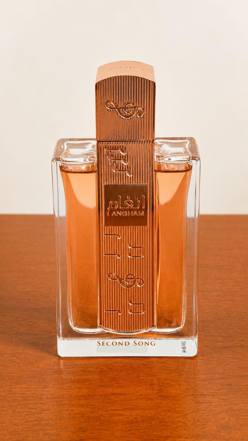 May include: A rectangular perfume bottle with a copper-colored cap and musical note accents. The bottle contains a light orange liquid and features the text "ANGHAM" in Arabic script. The base of the bottle reads "SECOND SONG".