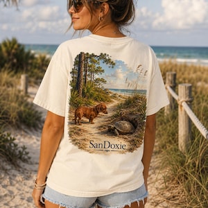 Puede incluir: Una camiseta de manga corta color hueso con un gráfico en la espalda de un perro salchicha marrón en un camino de arena junto a una tortuga, con pinos y vista al mar de fondo y el texto SanDoxie impreso en la parte inferior.