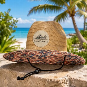 May include: A straw hat with a wide brim featuring a red, blue, and white patterned fabric underside. The front of the hat displays a circular patch with a graphic of a dachshund surfing a wave and the text SANDOXIE. A black chin strap is attached.