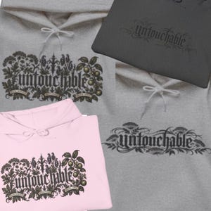Puede incluir: Una colección de sudaderas con capucha en gris claro, gris oscuro y rosa claro. Cada sudadera presenta un estampado gráfico con la palabra "untouchable" en una fuente gótica, rodeada de ilustraciones botánicas detalladas de plantas y frutas.