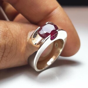 May include: A silver ring featuring a single oval-cut deep red ruby gemstone set into a wide, polished metal band. The band has a smooth, solid construction with a reflective surface that highlights the contrast between the metal and the stone.