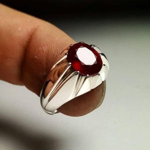 Könnte beinhalten: Ein silberner Ring mit einem großen, oval geschliffenen, tiefroten Edelstein in der Mitte. Die Ringschiene hat eine polierte metallische Oberfläche mit markanten, kantigen Rillen, die vom zentralen Steinbesatz zu den Seiten verlaufen.