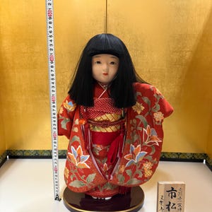 May include: A Japanese Ichimatsu doll with black hair, wearing a red kimono with a floral pattern and a gold obi. A measuring tape shows the doll is approximately 19.5 inches tall. A wooden plaque with Japanese text stands next to the doll.