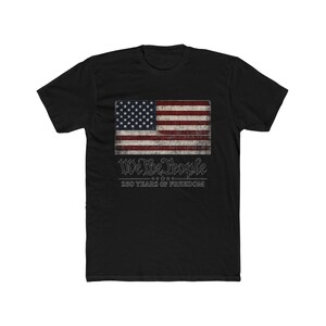 Könnte beinhalten: Ein schwarzes Kurzarm-T-Shirt mit einer Grafik der amerikanischen Flagge im Used-Look auf der Brust. Unter der Flagge steht der Text We The People, gefolgt von fünf Sternen und dem Schriftzug 250 YEARS OF FREEDOM in weißer Schrift.