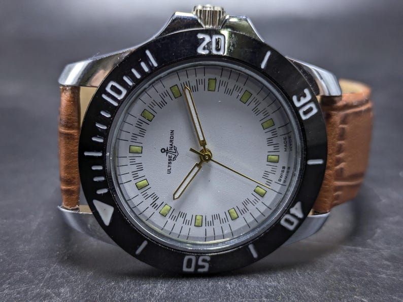 Może przedstawiać: Zegarek na rękę Ulysse Nardin ze srebrną tarczą, świecącymi indeksami godzin, złotymi wskaz&oacute;wkami i czarnym obrotowym pierścieniem z białymi cyframi. Zegarek posiada teksturowany jasnobrązowy sk&oacute;rzany pasek oraz srebrną kopertę.