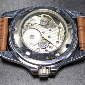 Może przedstawiać: Srebrzysty zegarek na rękę z przezroczystym deklem ukazującym wewnętrzny mechanizm. Na mechanizmie wygrawerowano napisy Swiss, seventeen, 17 jewels oraz Ulysse Nardin. Zegarek posiada jasnobrązowy sk&oacute;rzany pasek o wyraźnej fakturze.