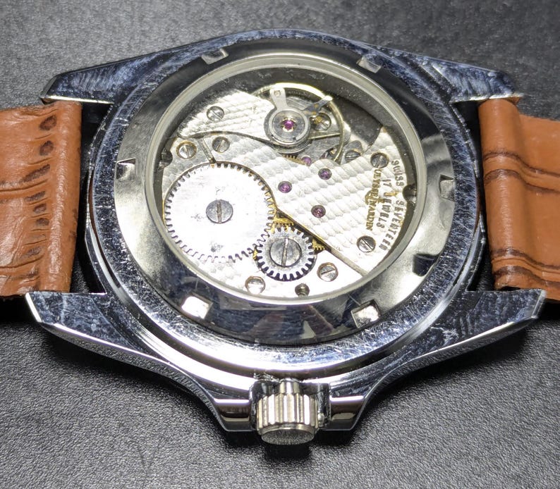 Może przedstawiać: Srebrzysty zegarek na rękę z przezroczystym deklem ukazującym wewnętrzny mechanizm. Na mechanizmie wygrawerowano napisy Swiss, seventeen, 17 jewels oraz Ulysse Nardin. Zegarek posiada jasnobrązowy sk&oacute;rzany pasek o wyraźnej fakturze.