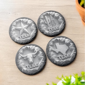 May include: Four circular slate coasters with rough, uneven edges. Each coaster features a raised, embossed design in the center: a five-pointed star, a cactus and oil derrick, a longhorn steer head, and the state map of Texas, all with the word TEXAS.