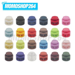 May include: A collection of twenty-four plastic cord locks arranged in a grid, featuring a variety of solid colors including white, black, blue, red, pink, yellow, green, brown, and clear. Each toggle has a spring-loaded button mechanism.