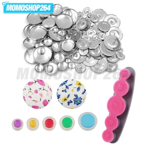May include: A collection of silver-coloured metal button components, two fabric-covered buttons with floral patterns, five colourful plastic buttons, and a pink and grey plastic measuring tool with circular cutouts labelled MOMOSHOP264.