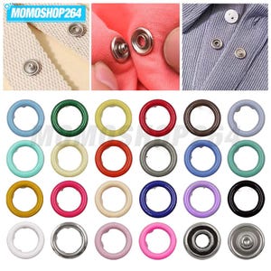 Pode incluir: Uma coleção de botões de pressão metálicos em várias cores, incluindo azul, verde, amarelo, vermelho, castanho, roxo e preto. O conjunto inclui anéis circulares e botões prateados fixados em tecidos de tons bege, rosa e azul.
