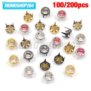 Può includere: Una collezione di borchie rotonde in metallo con griffe, dotate di cristalli sfaccettati trasparenti e rosa su basi color oro, argento e metallo scuro. Il testo MOMOSHOP264 e 100/200pcs è visibile. Queste chiusure hanno quattro griffe.