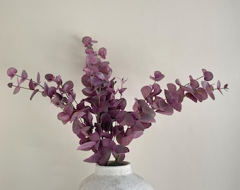 Op de afbeelding: Een witte, getextureerde, bolvormige vaas bevat verschillende stelen kunstmatige eucalyptus met afgeronde, stoffig paarse bladeren. De stelen vertakken zich in meerdere richtingen, wat zorgt voor een vol en symmetrisch arrangement van bladeren.