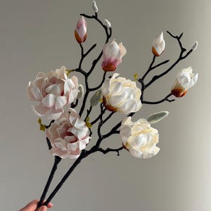 Könnte beinhalten: Ein Strauß künstlicher Magnolienblüten. Die Blüten befinden sich in verschiedenen Stadien der Blüte, einige als Knospen und andere vollständig geöffnet. Die Blütenblätter sind zartrosa und cremefarben, und die Stiele sind schwarz. Der Strauß wird in einer Hand gehalten.