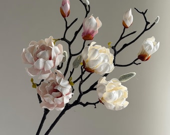 May include: A decorative branch featuring multiple magnolia flowers in shades of soft pink and creamy white. The branch is dark brown with a textured surface, accented by small, fuzzy, light green buds distributed along the slender, branching stems.