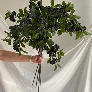 May include: A large artificial bouquet featuring multiple stems with dark blue, round berries and vibrant green leaves. The stems are gathered together at the base, showcasing a dense arrangement of realistic foliage and clusters of small, dark fruit.