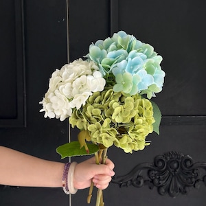 May include: A bouquet of artificial hydrangea flowers featuring three distinct clusters in shades of creamy white, soft pastel blue, and muted olive green. The stems are bundled together, showcasing detailed green leaves and rounded petal formations.