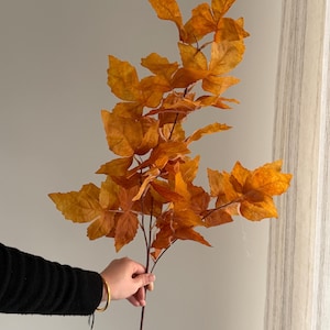 May include: A decorative artificial branch featuring multiple serrated leaves in shades of burnt orange and golden brown. The branch has a thin, dark brown stem that extends upward, supporting a cluster of textured, autumn-colored foliage.