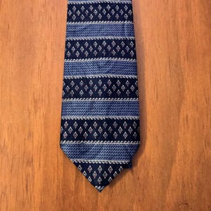 May include: A necktie featuring a repeating pattern of dark blue and light blue horizontal bands. The design includes rows of small white diamonds with red centers alternating with textured light blue sections and thin white scalloped lines.