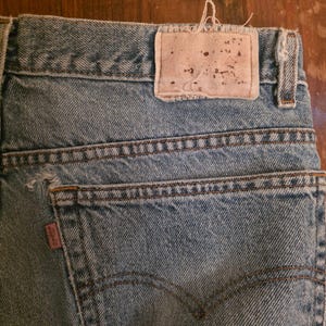 May include: Light blue denim jeans featuring a rectangular beige leather patch on the back waistband, a small red fabric tag with the Levi's brand name on the back pocket, and visible stitching details along the pockets and seams of the garment.