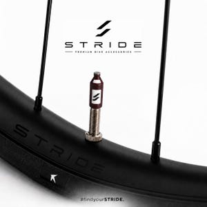 Könnte beinhalten: Eine dunkel-kastanienbraune und weiße Ventilkappe in Form einer kleinen Flasche mit dem Stride-Logo, befestigt an einem Metall-Fahrradventil an einer schwarzen Felge. Der Text STRIDE PREMIUM BIKE ACCESSORIES und #findyourSTRIDE. sind sichtbar.