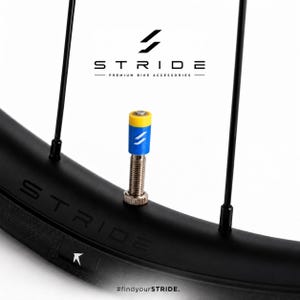 Könnte beinhalten: Eine Fahrradventil-Kappe mit gelbem Oberteil und blauem Unterteil mit weißem Logo, befestigt an einem silbernen Metall-Ventilschaft auf einer schwarzen Felge. Der Text STRIDE PREMIUM BIKE ACCESSORIES und #findyourSTRIDE sind im Bild zu sehen.