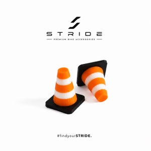 Könnte beinhalten: Zwei Miniatur-Verkehrskegel mit orangefarbenen und weißen horizontalen Streifen auf schwarzen quadratischen Sockeln. Über den Kegeln steht STRIDE PREMIUM BIKE ACCESSORIES. Darunter steht der Text #findyourSTRIDE.