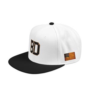 May include: A white snapback cap with a black flat brim and a black button on top. The front features the letters BD in bold black font with a gold outline. On the side, there is a small rectangular patch displaying the American flag in red, white, and blue.