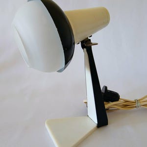Może przedstawiać: Zabytkowa lampa biurkowa w kolorach białym i czarnym. Lampa ma trójkątną podstawę, czarno-białe wsporniki i cylindryczną głowicę. Głowica lampy jest biała z czarnym akcentem. Lampa ma przewód.