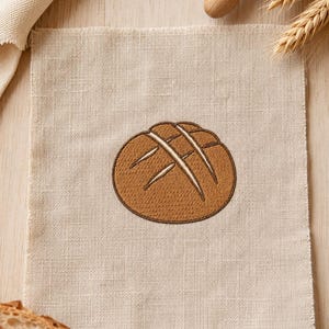 May include: A square piece of light beige fabric featuring a centered machine embroidery design of a round, golden-brown loaf of bread with three distinct diagonal score lines across the top surface, rendered in a simple, clean graphic style.