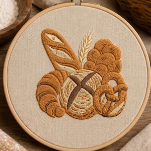 May include: A circular wooden embroidery hoop containing a beige fabric panel with a detailed thread-stitched design of various baked goods, including a baguette, a round loaf, a croissant, a pretzel, and a stalk of wheat in shades of brown.