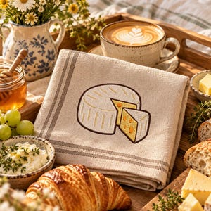 Peut inclure: Une serviette en lin beige pliée ornée d'une broderie représentant une meule de fromage ronde avec une part découpée. La serviette présente deux fines rayures brunes sur le bord. La broderie montre une croûte blanche et un intérieur jaune.