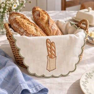 Puede incluir: Una cesta de mimbre tejida con un forro de tela color crema con bordes festoneados que presenta un bordado de dos baguettes en una bolsa de papel. Dos hogazas de pan crujiente descansan dentro de la cesta de acabado natural marrón claro.