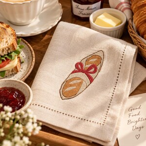 Puede incluir: Una servilleta de lino beige con una baguette bordada atada con una cinta roja. La servilleta tiene un borde con dobladillo de vainica y está colocada sobre una bandeja de madera junto a café, mermelada, mantequilla y un croissant.
