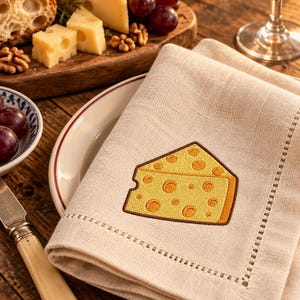 May include: A beige linen napkin featuring an embroidered yellow wedge of Swiss cheese with brown outlines. The napkin has a hemstitched border and is folded neatly on a white plate with a thin brown rim, showcasing a detailed fabric texture.