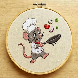 Könnte beinhalten: Gesticktes Kunstwerk in einem Holzreifen zeigt eine Cartoon-Chefmaus mit weißer Kochmütze und -jacke, die eine Bratpfanne hält. Gemüse, darunter eine Tomate, ein Pilz und eine Zwiebel, werden als geworfen dargestellt. Die Maus wirkt gestresst.