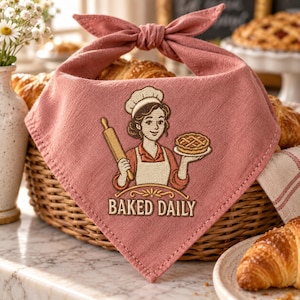 May include: A dusty rose triangular pet bandana featuring an embroidered illustration of a baker wearing a chef hat and apron, holding a rolling pin and a pie. The text "BAKED DAILY" is embroidered in cream below the figure. The fabric has a tie top.