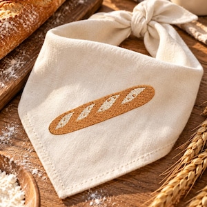 May include: A cream-colored fabric bandana featuring an embroidered golden-brown baguette design with white diagonal slashes. The triangular accessory is tied in a knot at the top, showcasing a clean hemmed edge and a textured woven material.