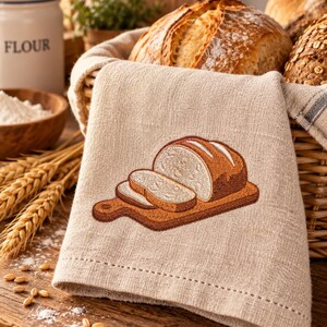 Op de afbeelding: Een lichtbeige linnen theedoek met een geborduurd ontwerp van een gesneden brood op een houten snijplank. Het borduurwerk gebruikt tinten bruin en crème om het brood en de plank in het midden van het stoffen oppervlak weer te geven.