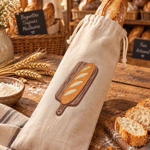 Könnte beinhalten: Ein beigefarbener Brotsack aus Leinen mit Kordelzug, der mit einer Stickerei eines Baguettes auf einem Holzschneidebrett versehen ist. Der Sack ist mit einem knusprigen Baguette gefüllt, dessen oberes Ende aus der Stofföffnung herausragt.
