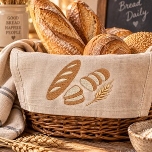 Può includere: Un cesto rotondo in vimini intrecciato foderato con un panno di lino beige caratterizzato da ricami di una baguette, pane a fette e una spiga di grano. Il cesto è riempito con vari tipi di pane croccante, tra cui una baguette e panini.
