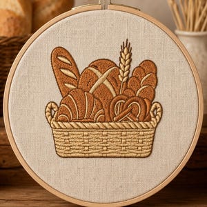 May include: A circular wooden embroidery hoop framing a piece of light beige fabric. The fabric features a detailed embroidery design of a woven basket filled with various types of brown bread, including a baguette, a pretzel, and a stalk of wheat.